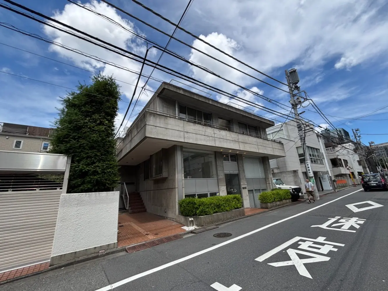 - コンクリート打ちっぱなしのモダンな建物、青空と白い雲、街路の植物と電線が見える風景