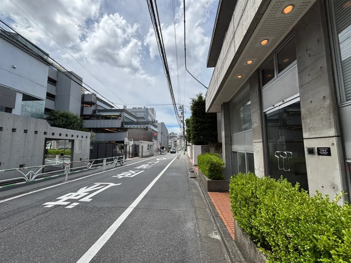 - コンクリートの建物とガラスのエントランスが見える街の風景、片側に緑の植栽と広がる道路、青空と白い雲の背景