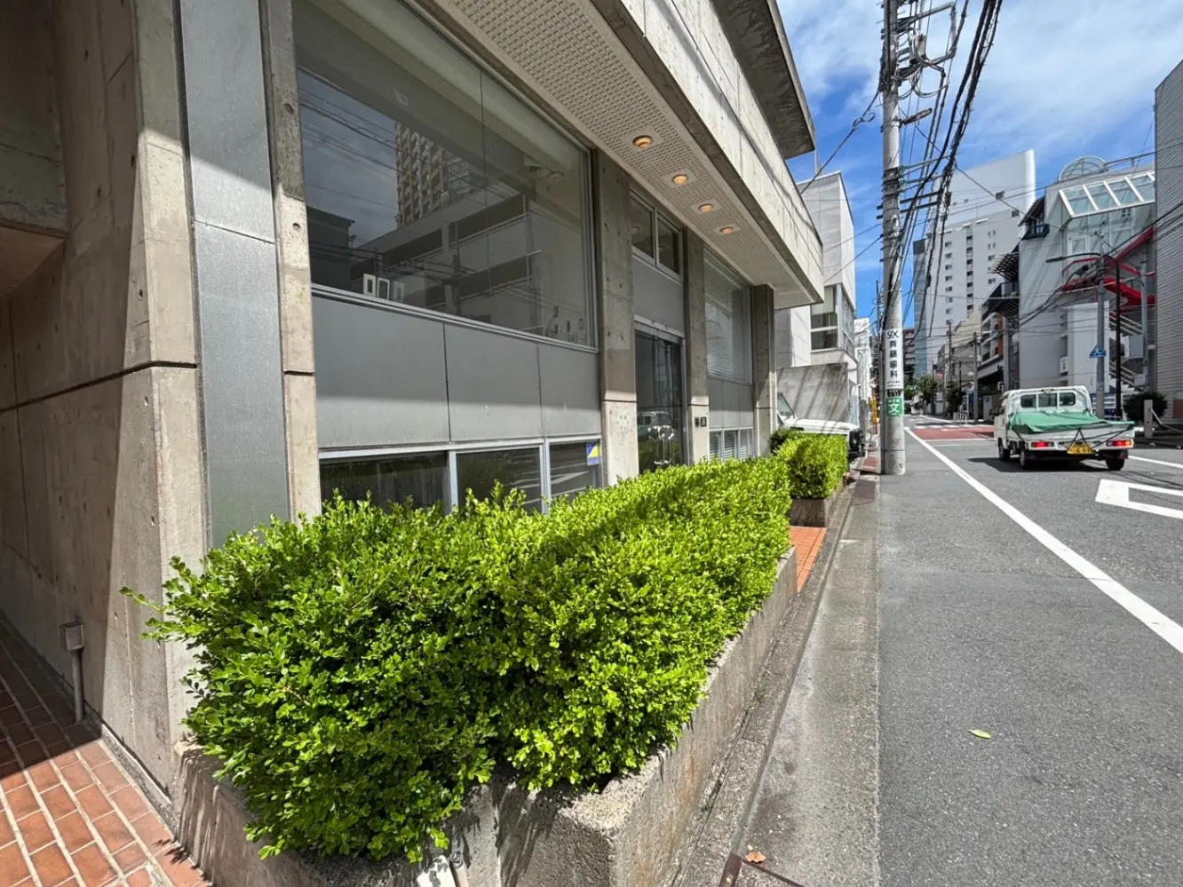 - 街中のコンクリート造りの建物と緑の生垣、青空と電柱が見える通りの風景