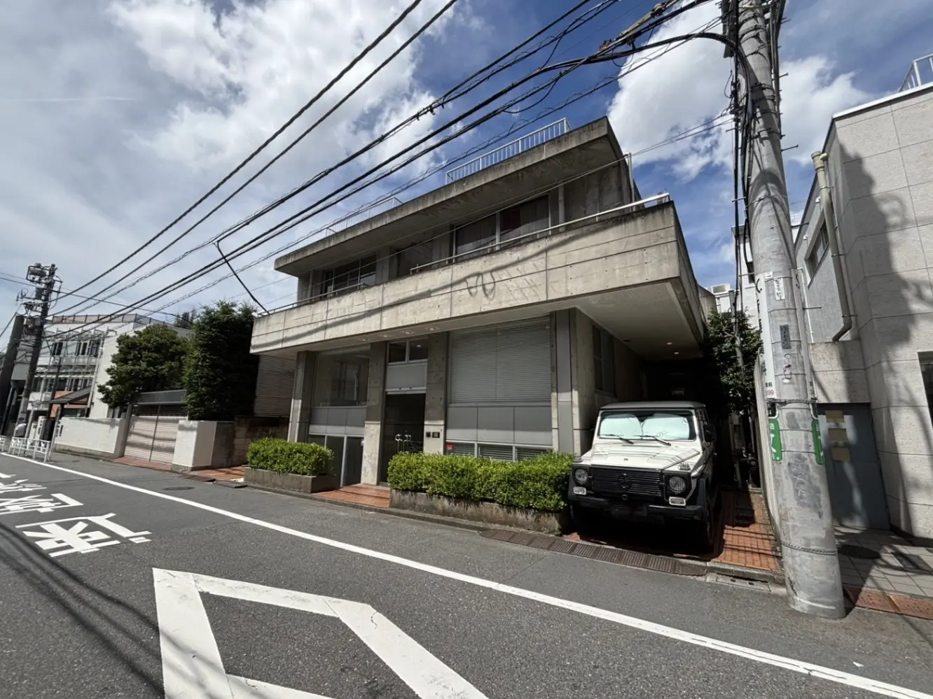 - 近代的なコンクリートの建物とその前に駐車された白いSUV、周囲に豊かな緑と電柱が立ち並ぶ街並み