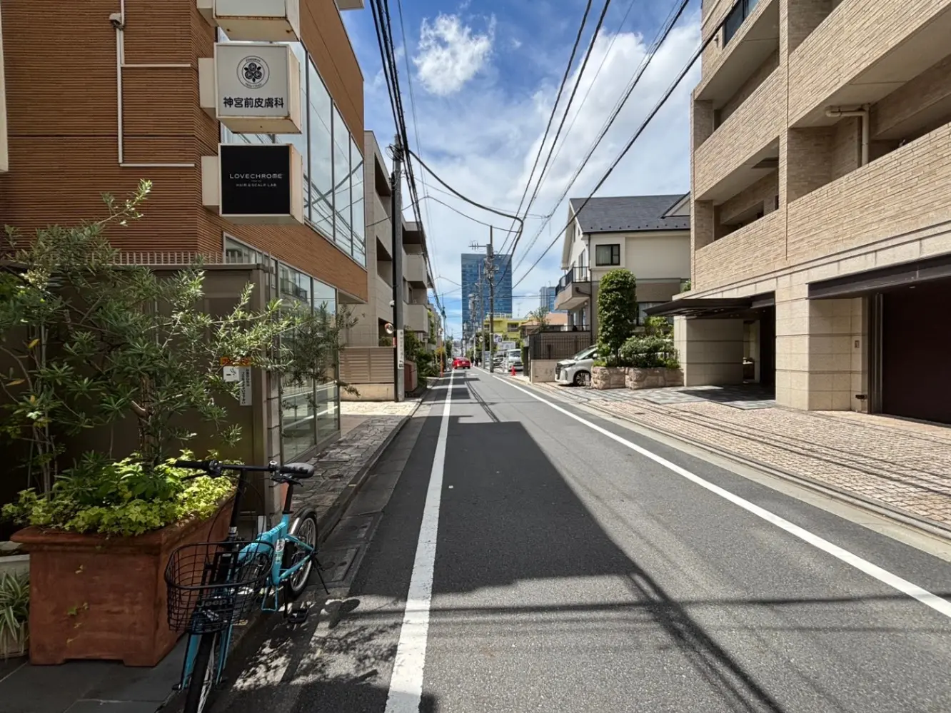 - 青い自転車が置かれた街並み、近代的な建物と緑の植栽が調和した通りの風景、青空の下に伸びる電線が特徴的なシーン