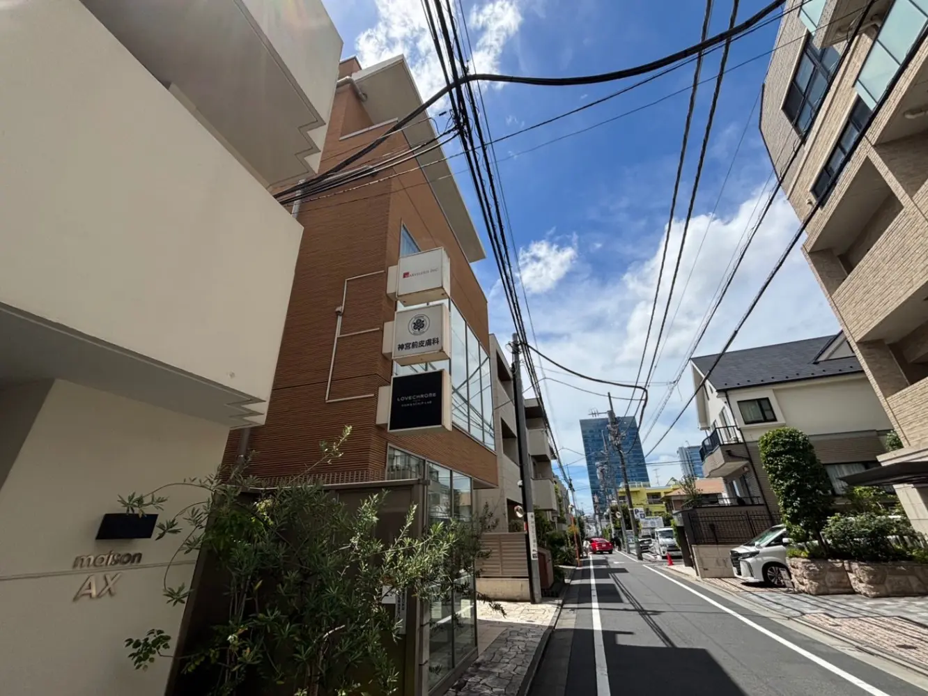 - 住宅街の通り沿いに立つモダンな建物、店舗看板や電線が見える風景、青空が広がる昼下がりの景色