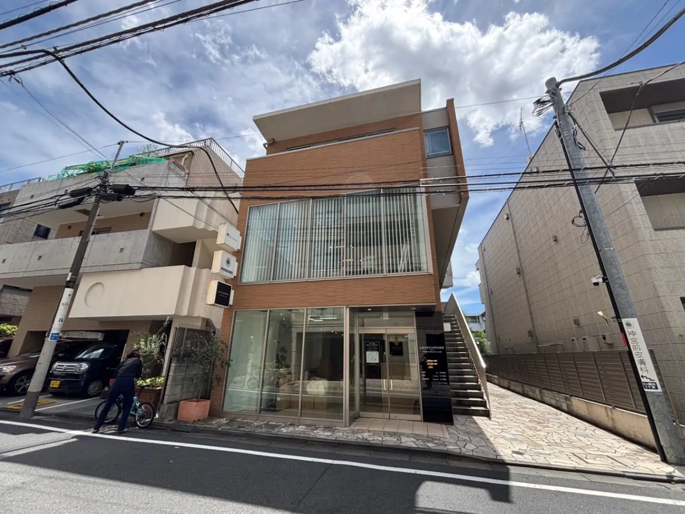 - 現代的な外観の商業ビル、広いガラス窓と木材のファサード、周囲の街並みと自転車利用者の姿が映える通りの風景