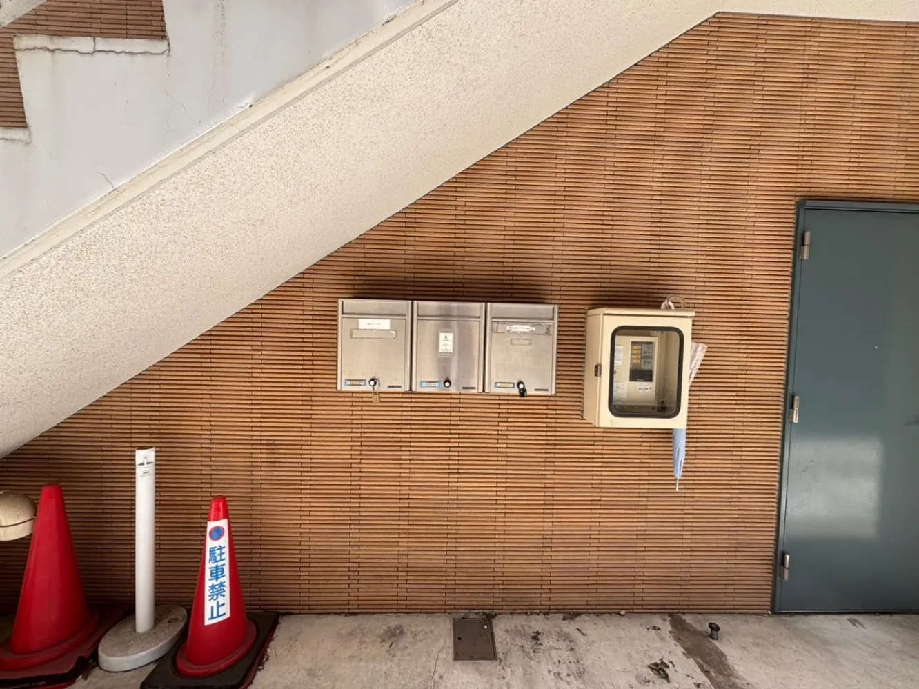 - 階段下に設置された郵便受けと電話ボックス、背景に木目調の壁、左側にある駐車禁止のコーンが目立つ風景