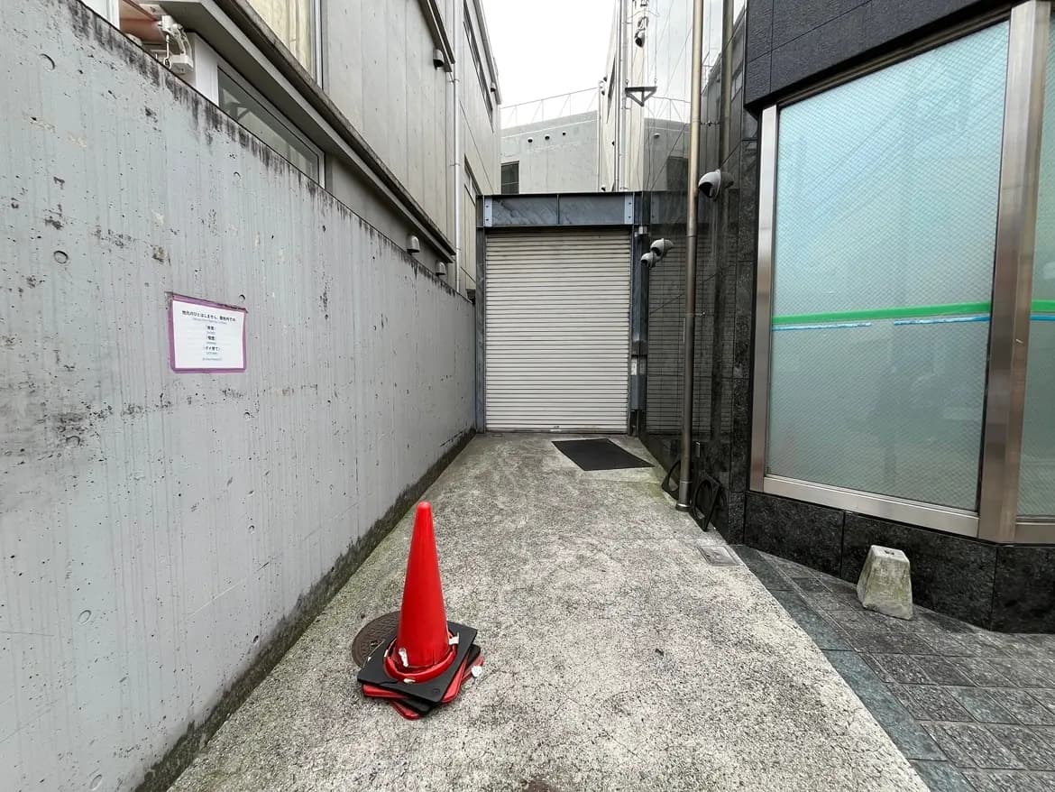 - シャッター前の狭い通路、赤いコーンと壁のコンクリート打ちっぱなし、近くにセキュリティカメラが設置された現代的なビルの一角