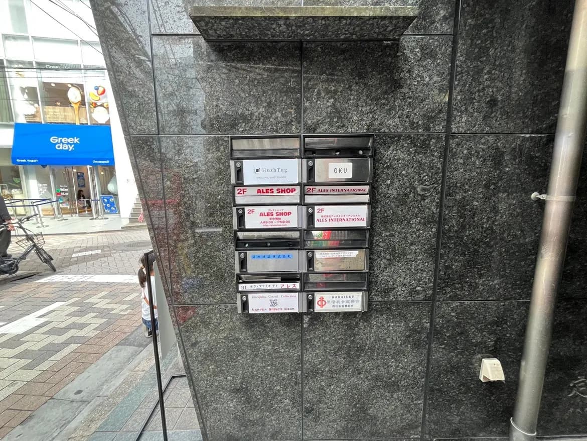 - グレーの石壁に取り付けられた複数の郵便受け、周囲には商業施設の入口と自転車が見える街の風景