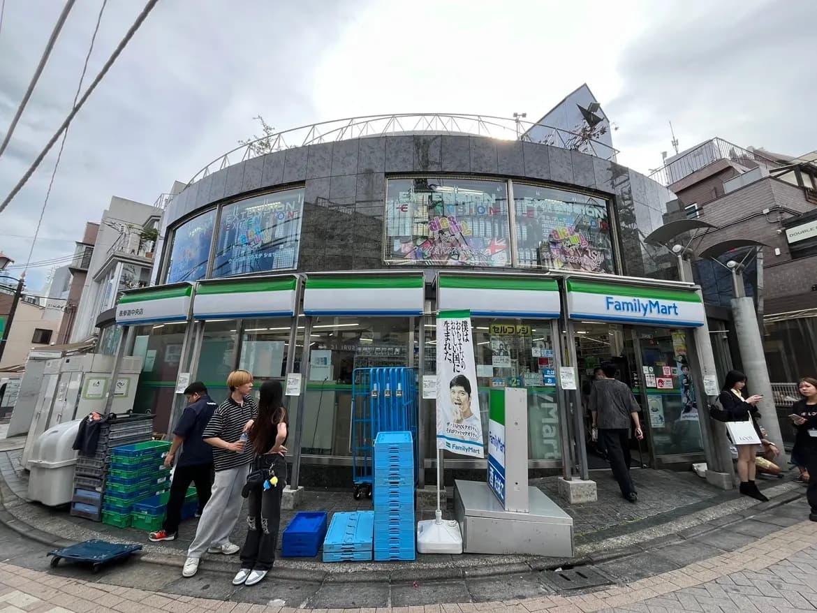 ファミリーマートの店舗外観、ガラス窓にカラフルなポスター、近くに人々が行き交う賑やかな印象の街角シーン