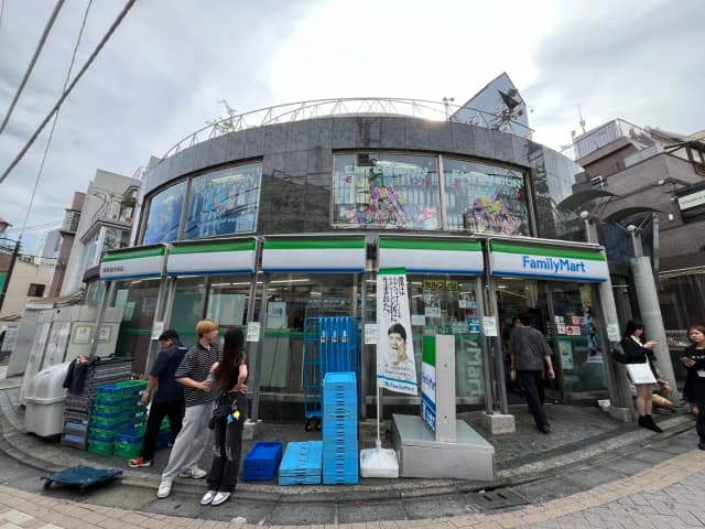 ファミリーマートの店舗外観、ガラス窓にカラフルなポスター、近くに人々が行き交う賑やかな印象の街角シーン