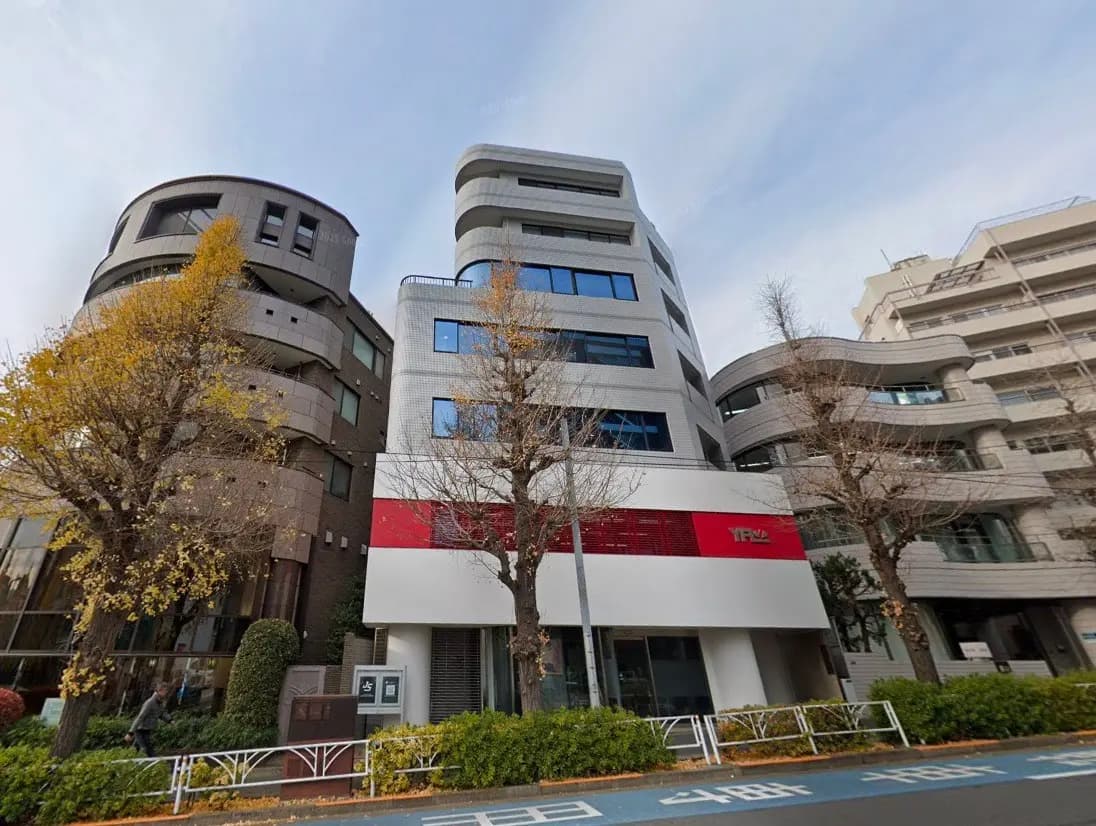 - モダンな外観のオフィスビル、周囲には黄葉した木々と整然とした街並み、エントランスには赤いアクセントが施されたデザイン