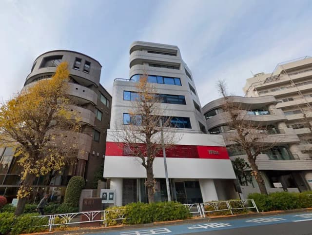 - モダンな外観のオフィスビル、周囲には黄葉した木々と整然とした街並み、エントランスには赤いアクセントが施されたデザイン