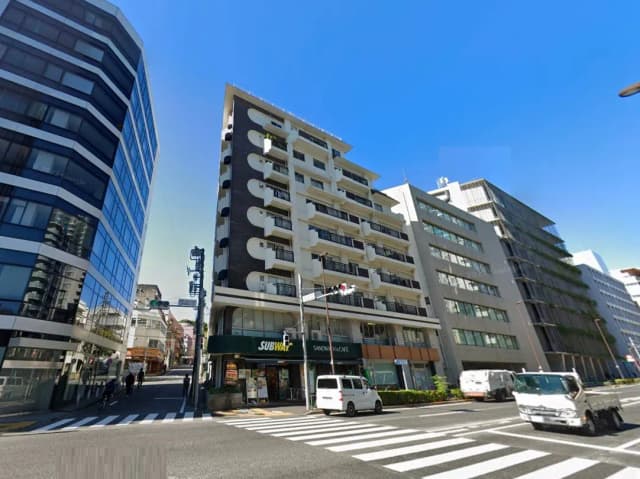 - 都市部の交差点に面した商業ビル群、ガラス張りのビルとバルコニーのある住宅ビルが並ぶ風景、青空が広がる日中の景色