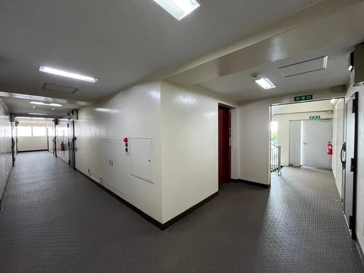 - 明るい照明のあるマンションの廊下、白い壁と整然とした床、非常口とエレベーターのドアが見える空間