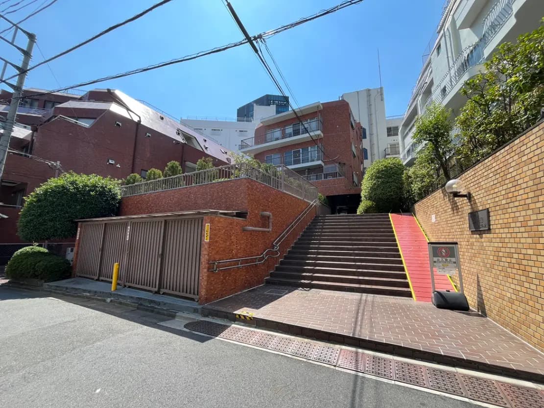 - 明るい青空の下、レンガ造りの建物と緑の植栽がある階段の風景、手すり付きのスロープと近くの収納ボックスが見える