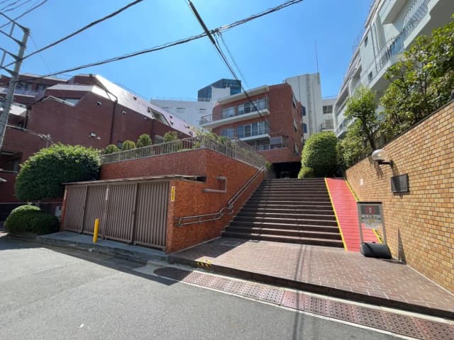 - 明るい青空の下、レンガ造りの建物と緑の植栽がある階段の風景、手すり付きのスロープと近くの収納ボックスが見える