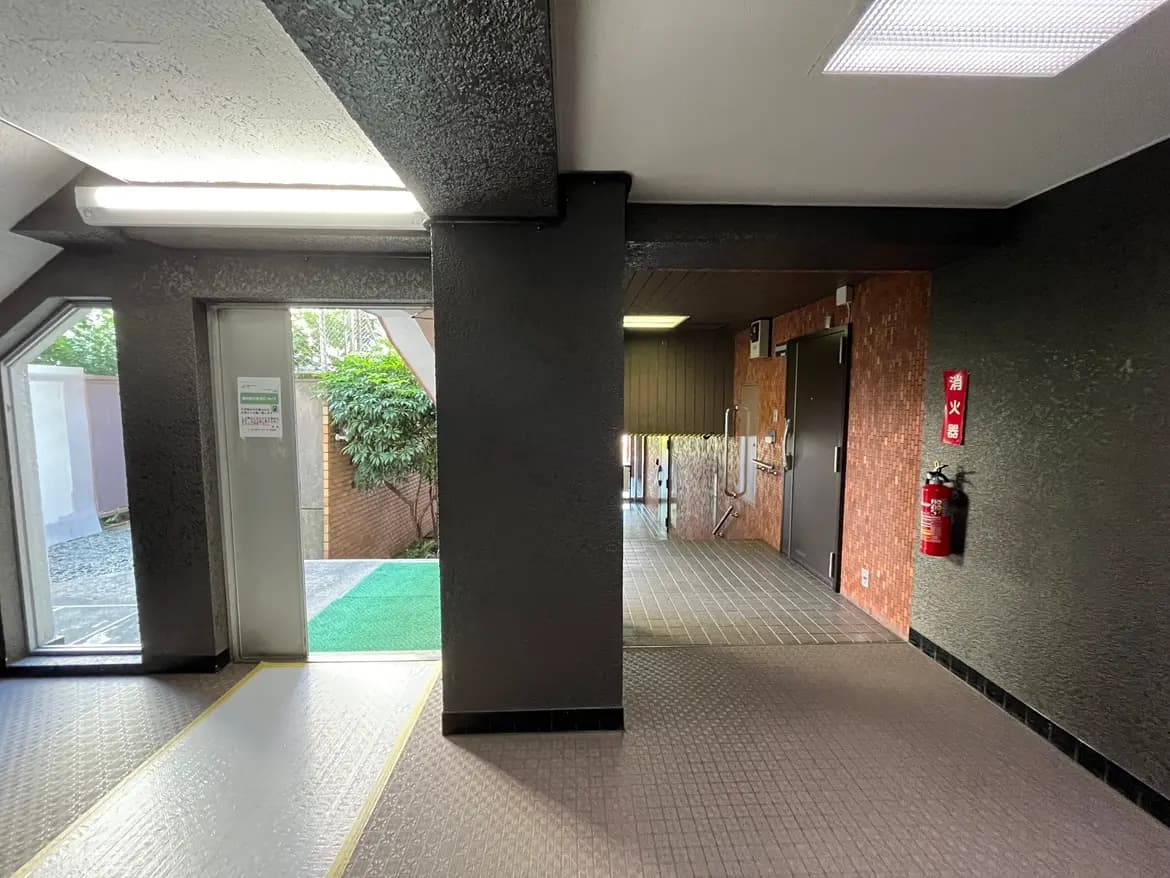 - モダンなデザインのエントランスホール、照明付きの廊下と防火設備、外に続く扉と緑の植栽が見える風景