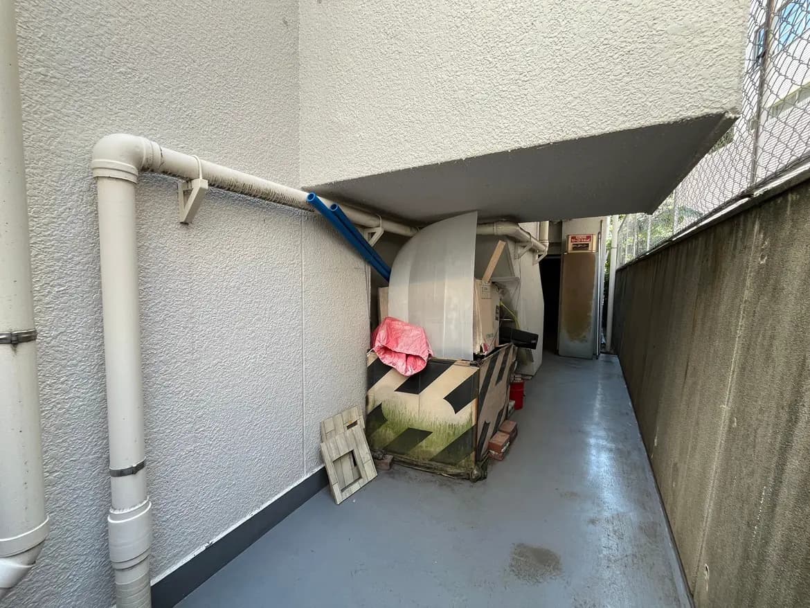 - 壁面に配管があり、物置きとして使用されている狭い通路、反対側にはフェンスが見える環境