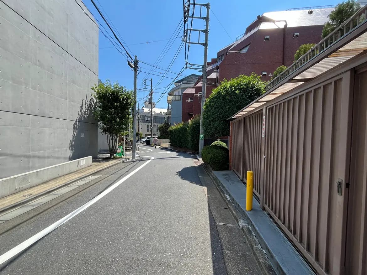 - 明るい青空の下、コンクリートの壁と並んだ住宅街の狭い街路、交通標識と緑の植栽が見える風景