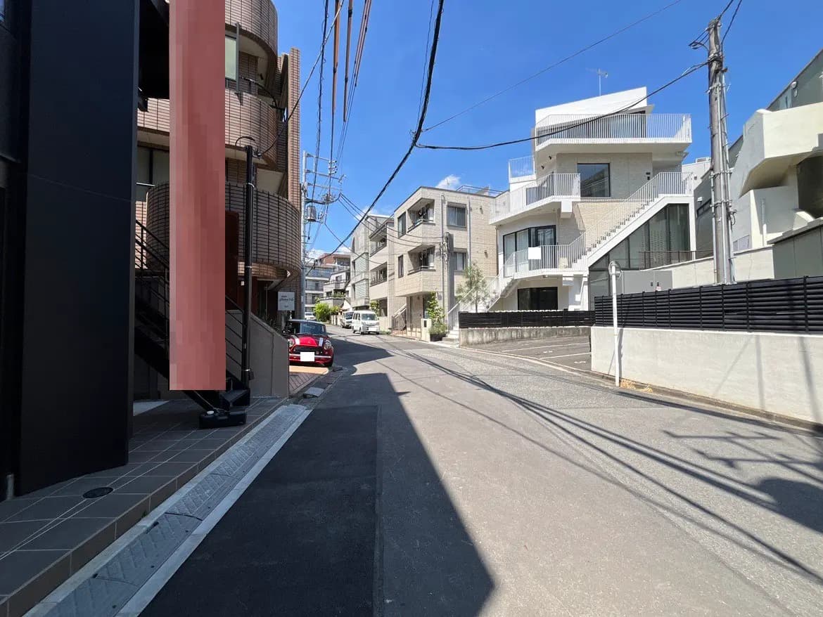 - 明るい青空の下、住宅街の静かな通りと現代的なデザインの建物が並ぶ風景、手前には階段と建物の外観が映るシーン
