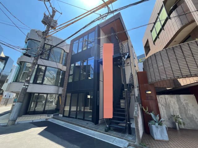 - 現代的な外観の3階建てビル、ガラスとタイルで構成されたファサード、青空の下に見える階段と電柱が並ぶ街の風景