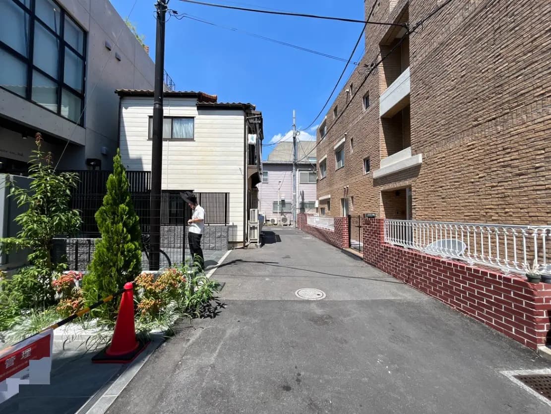 - 緑豊かな庭と赤い柵が特徴の路地、背景には複数の建物と青空が広がる風景