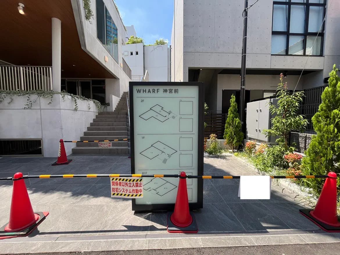 - WHARF神宮前の施設案内看板、階段と緑の植物に囲まれた現代的な建物の外観、交通規制用のコーンとバリケードが設置されている様子