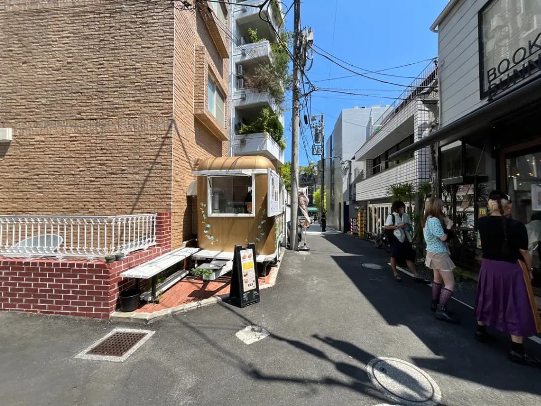 - 狭い路地に並ぶ飲食店とカフェのテント、外で待つお客さん、青空の下のカジュアルな雰囲気の街並み