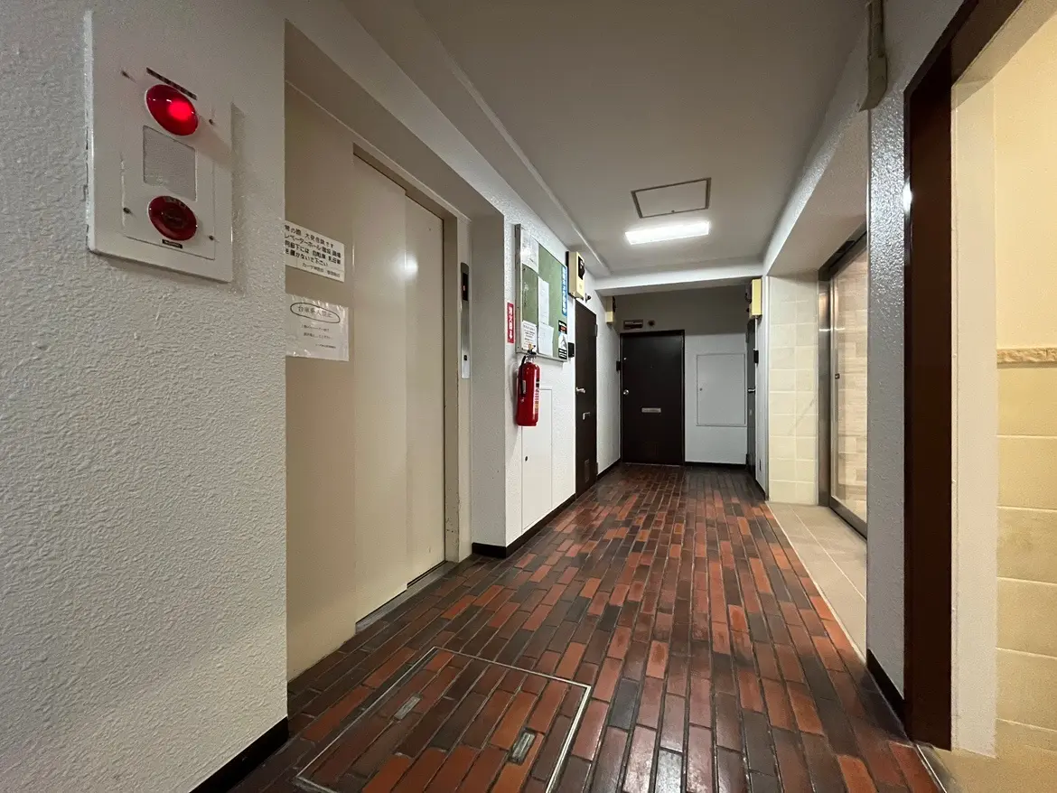 - 照明が明るいマンションのエントランスホール、エレベーターのボタンや掲示板、タイル張りの床が特徴の内装