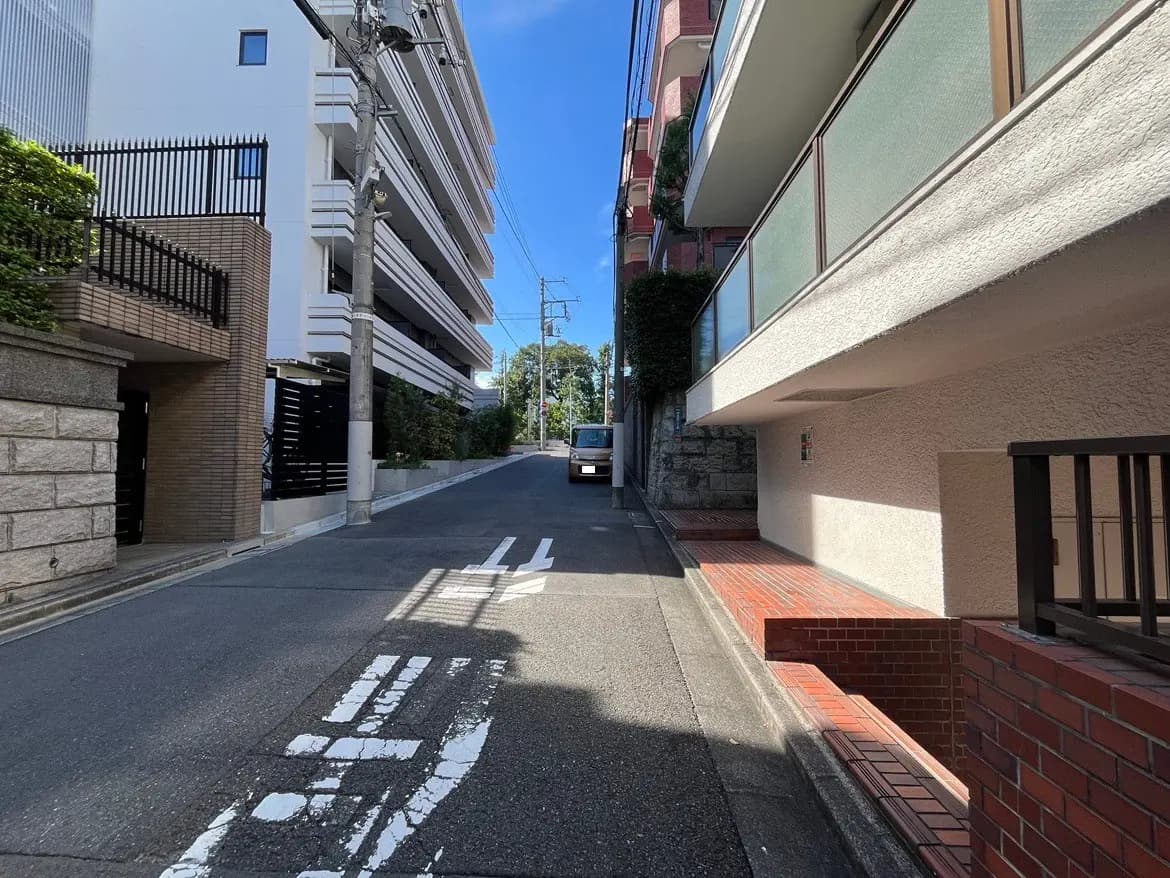 - 鮮やかな青空の下、両側に建物が並ぶ静かな住宅街の一角、舗装された道路とその先に見える車の風景