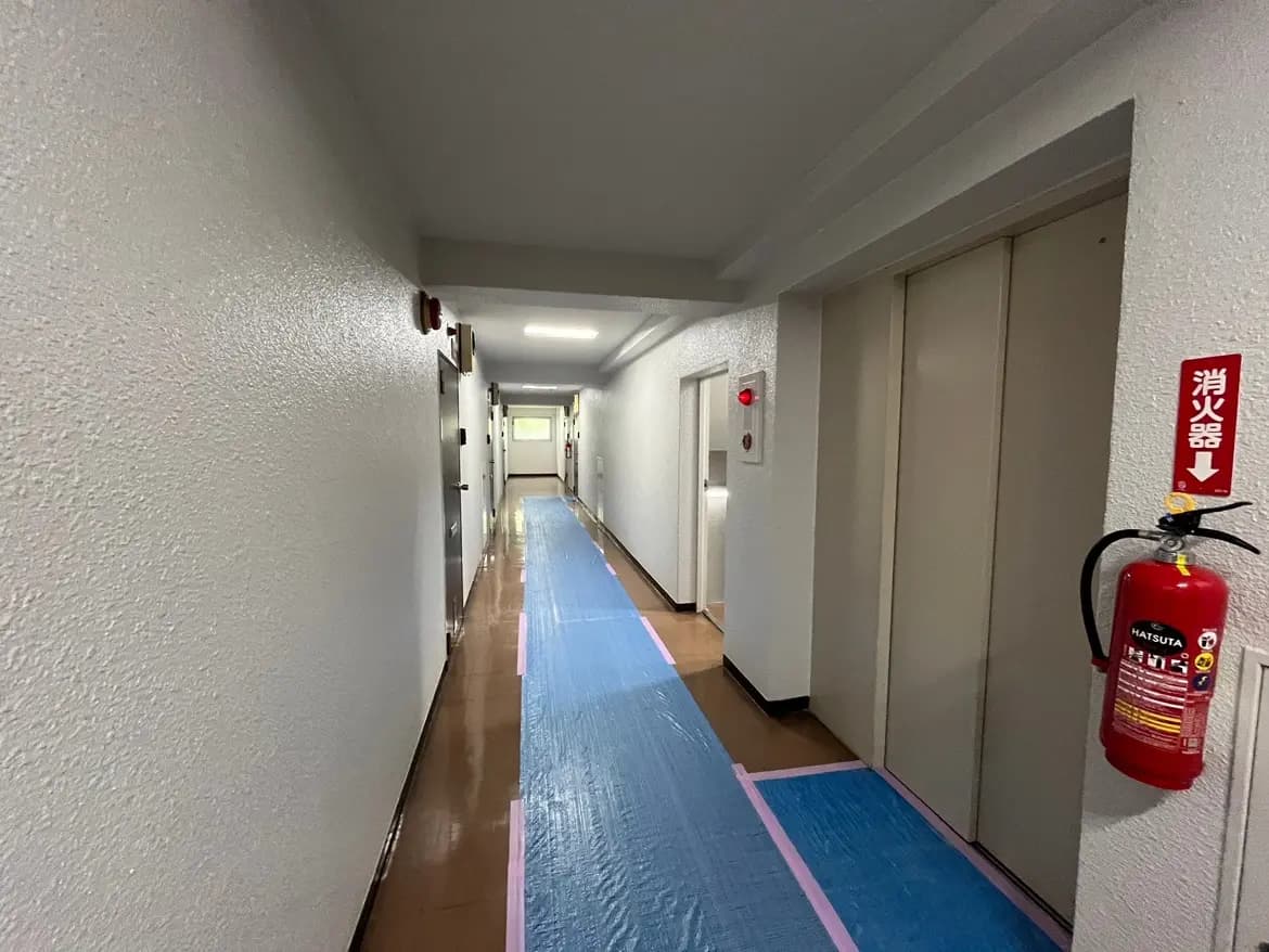 - 清潔感のあるホテルの廊下、白い壁と照明、青い保護シートが敷かれた床、エレベーターと消火器の設置された空間