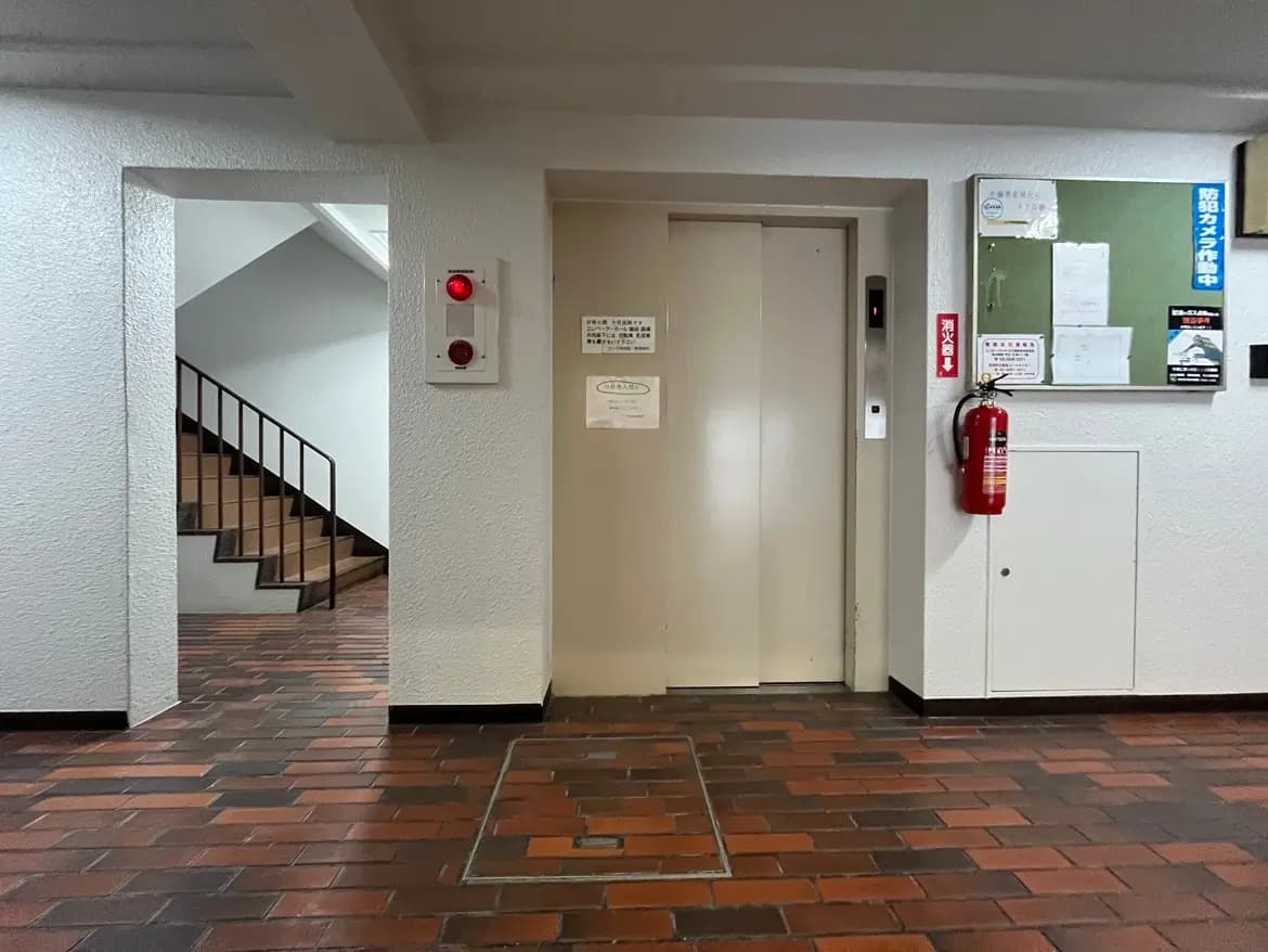 - エレベーターと階段のあるビルのロビー、対面に火災警報器と掲示板が設置された明るいスペース
