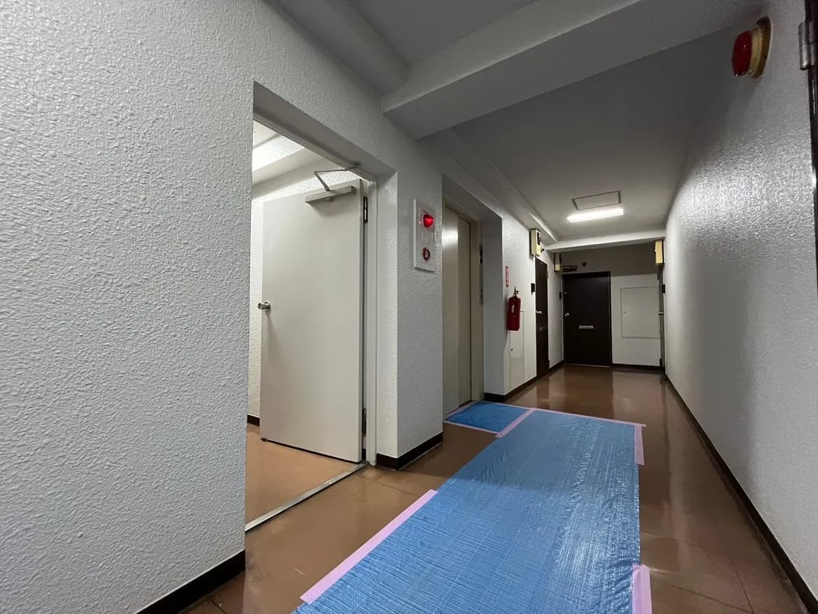 - 清潔感のある白い壁の廊下、青い養生シートが敷かれ、エレベーターといくつかのドアが並んでいる内部の景観