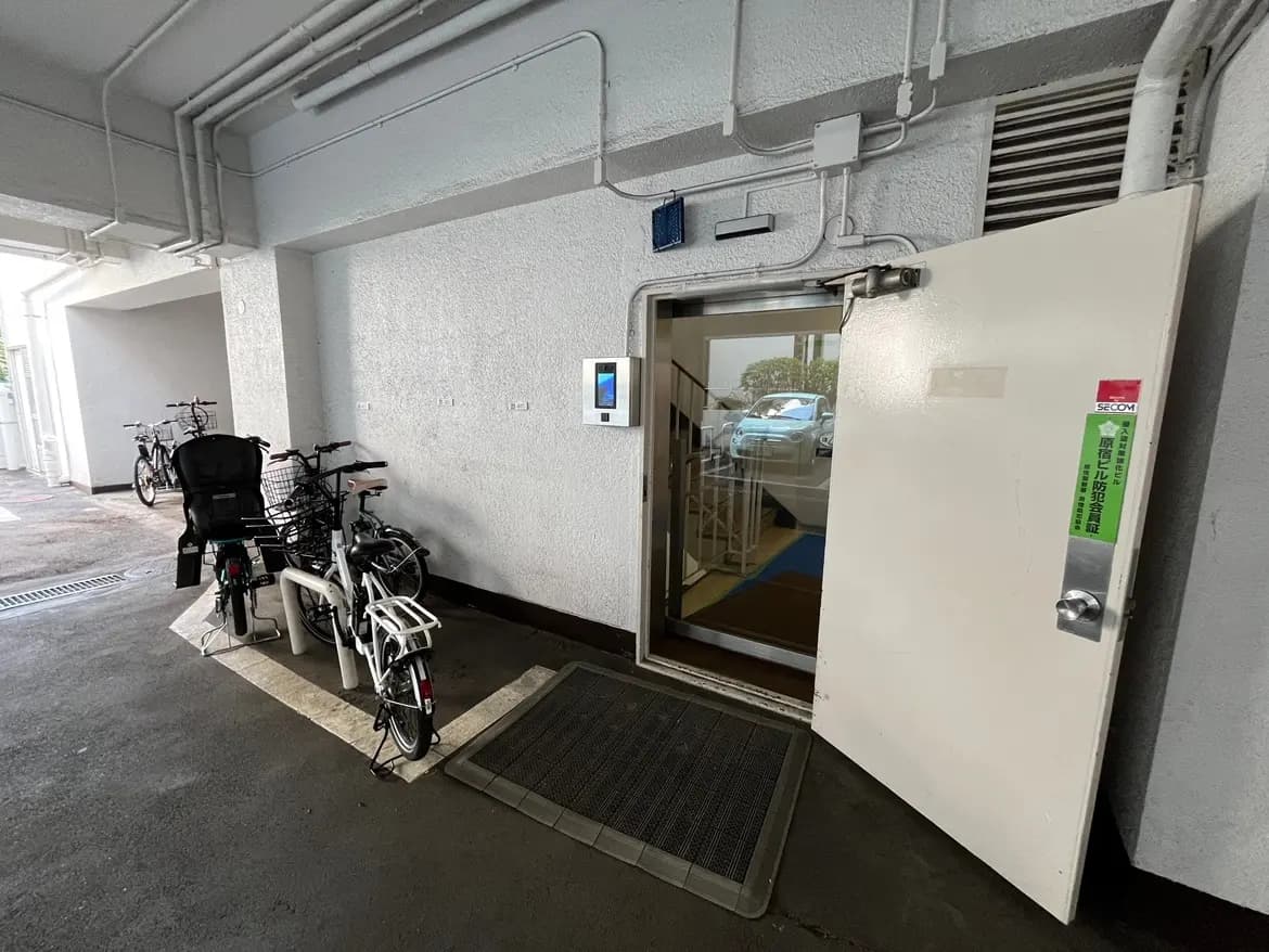 - 自転車置き場と防犯システムのあるエントランス、白い壁と開いたドア、屋内の様子が見える静かな環境