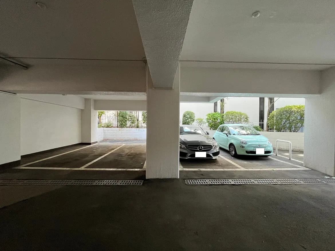 - 光が差し込む広々とした地下駐車場、白い壁と空いている駐車スペース、隣に停まる2台の車の様子
