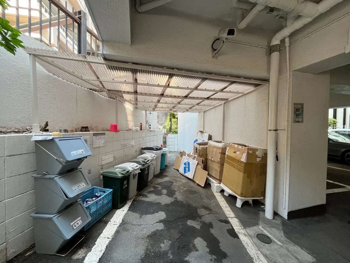 - ゴミ箱と段ボールが並べられた駐車場の隅、屋根付きの作業スペースと清掃用具が配置されたエリア
