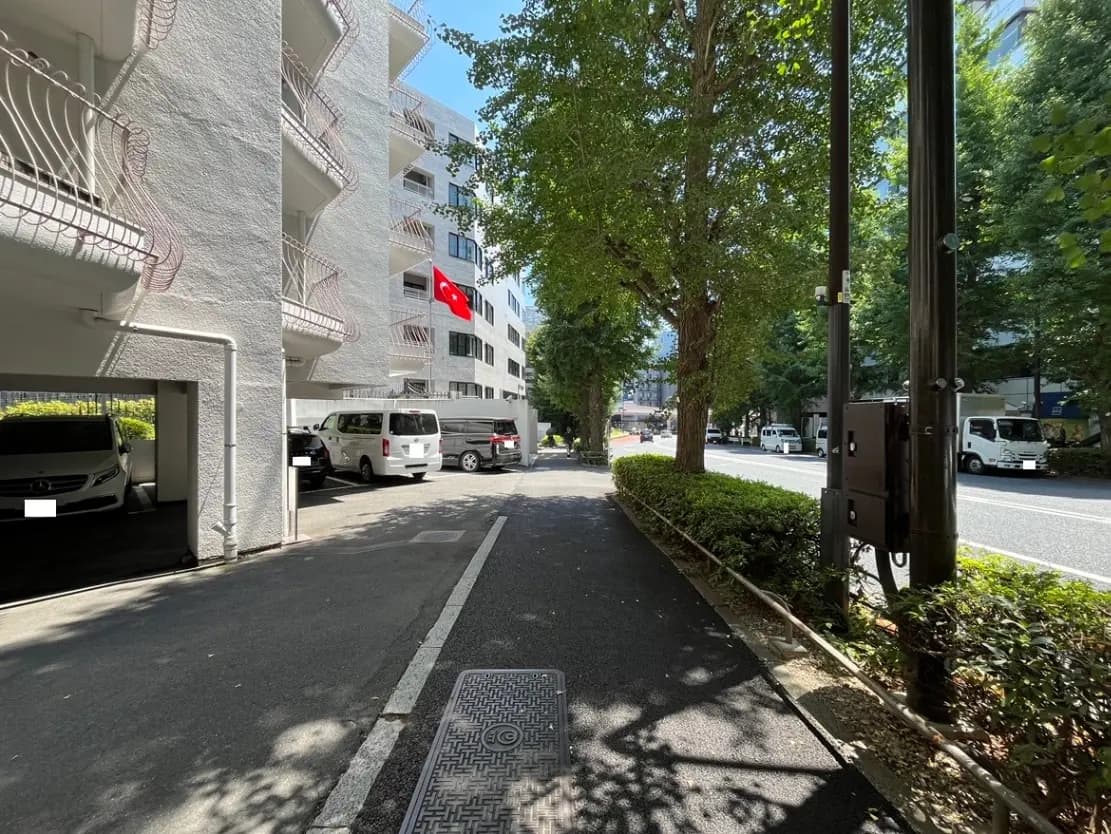 - 日差しの中、緑豊かな街路樹に囲まれた静かな住宅街の路地、隣接する建物の駐車場とトルコ国旗が目を引く風景