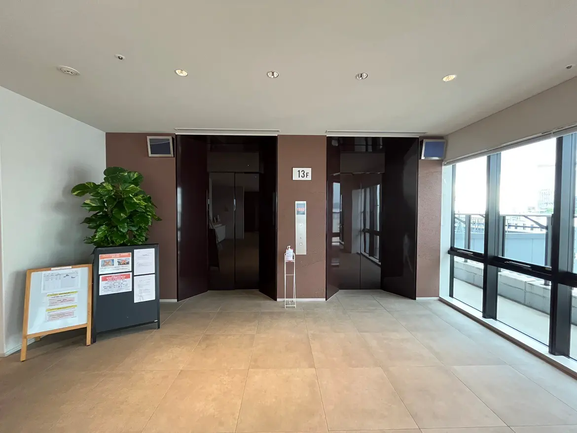 - 現代的なエレベーター前のエントランス、暖色系の壁、観葉植物と案内掲示板、広いガラス窓からの自然光が差し込む空間