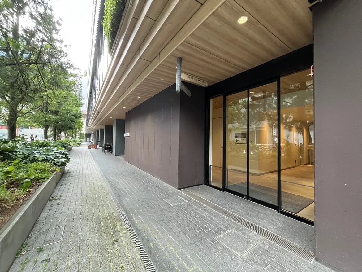 - 緑豊かな街路とモダンな建物のエントランス、ガラスのドアと木目調の天井が特徴の外観