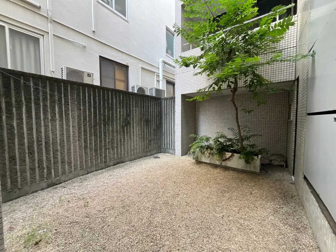 - 落ち着いた雰囲気の庭、砂利の敷かれたスペースと周囲には緑の植栽、隣にはビルの外壁が見える中庭の風景
