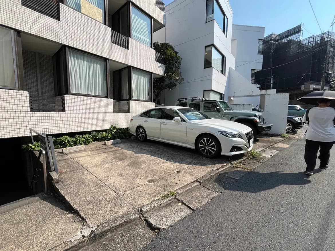 - 都市の住宅街にある白いセダンとSUVが駐車された駐車場、周囲の緑と近代的な建物が映る風景