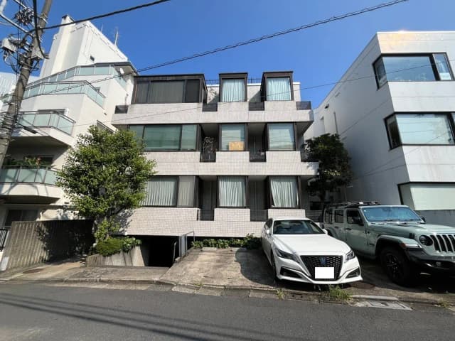 - モダンな外観のビル、白いタイル張りの壁と大きな窓、グリーンの植栽が施されたエントランスと隣接する車の駐車スペース