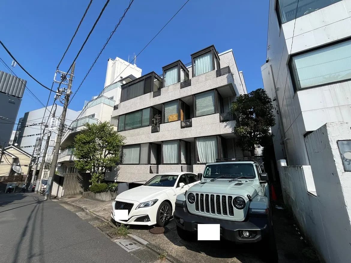 - 現代的な外観の住宅ビル、周囲に駐車された白と緑のSUV、青空が広がる街並みの風景