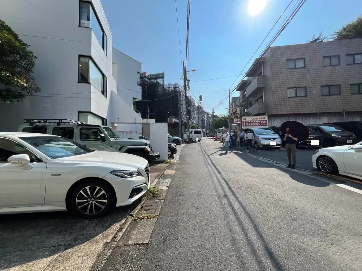 - 都市の道路沿いに停車した白い車と緑色の車両、周囲の建物や青空が広がる光景、駐車場の看板も見える。