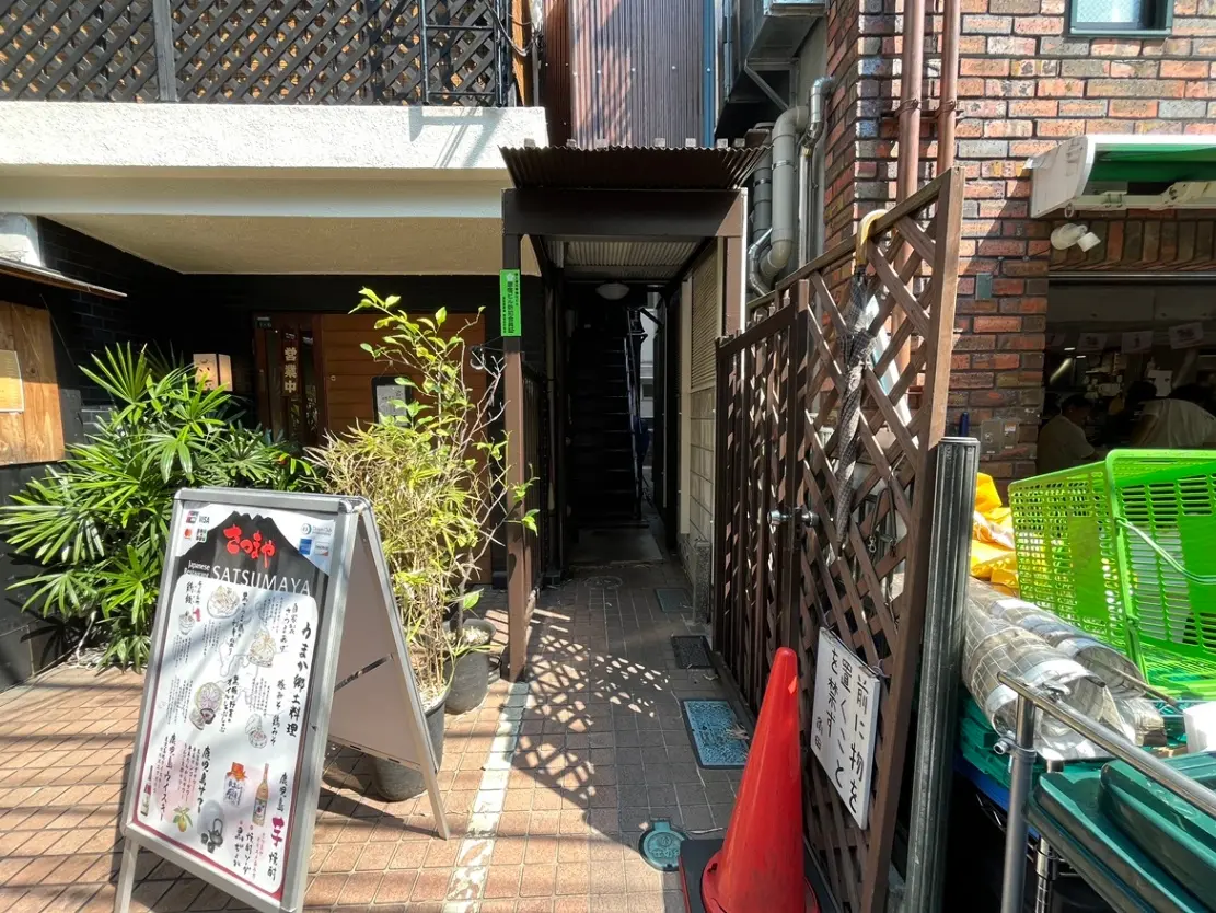 - 植物に囲まれた飲食店の入口、メニュー看板と通路が見える賑やかな商業エリアの風景