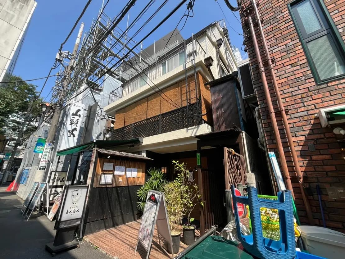- 外観が特徴的な和風居酒屋の入口、隣接するビルと青空が映える街並みの一角、看板と植栽が目を引くシーン