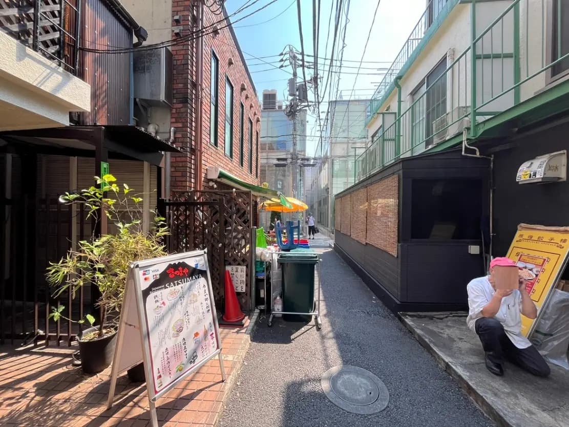 - 狭い路地にある飲食店の看板や屋台、赤い煉瓦の壁と青空の下でリラックスする店主の姿