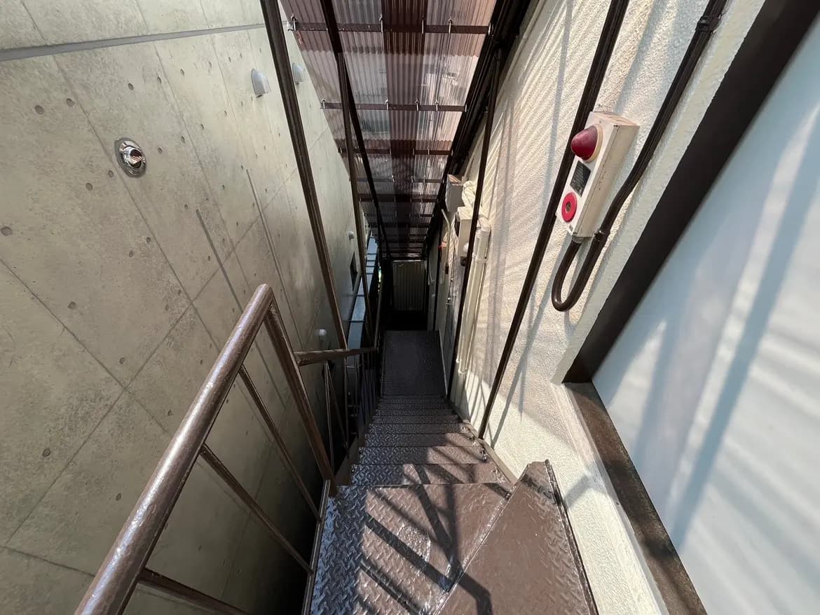 - 鉄製の手すりが付いた屋外の階段、コンクリートの壁と自然光が差し込む明るい通路の風景