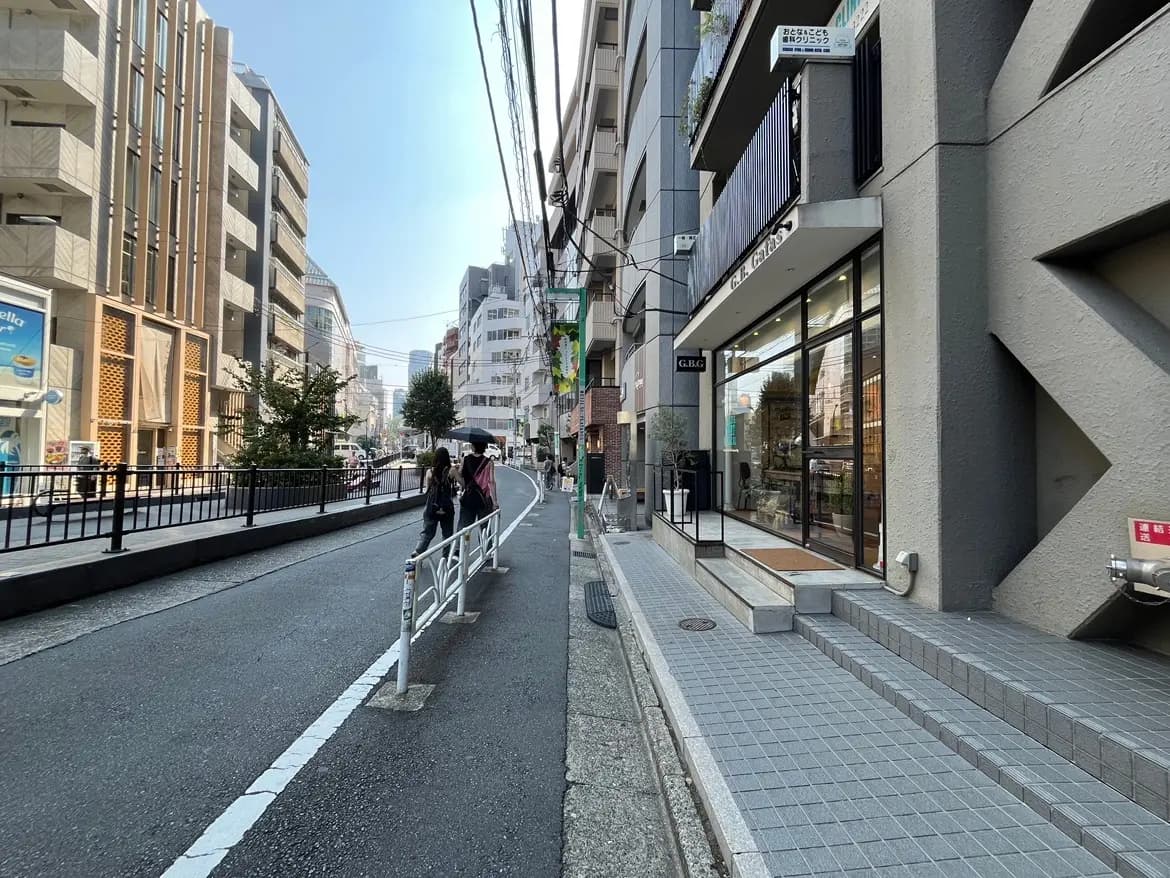 - 都市の通り沿いに並ぶ建物、歩道を歩く人々、晴れた空と店舗のガラス張りのファサードが特徴的な風景