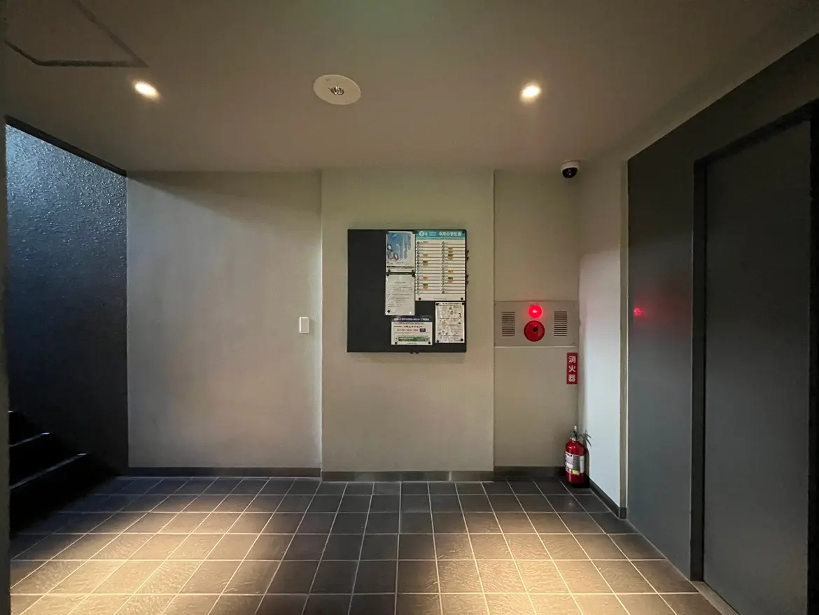 - モダンなデザインの建物のエントランス、掲示板、消防設備、暗めの照明とタイルフロアの内装