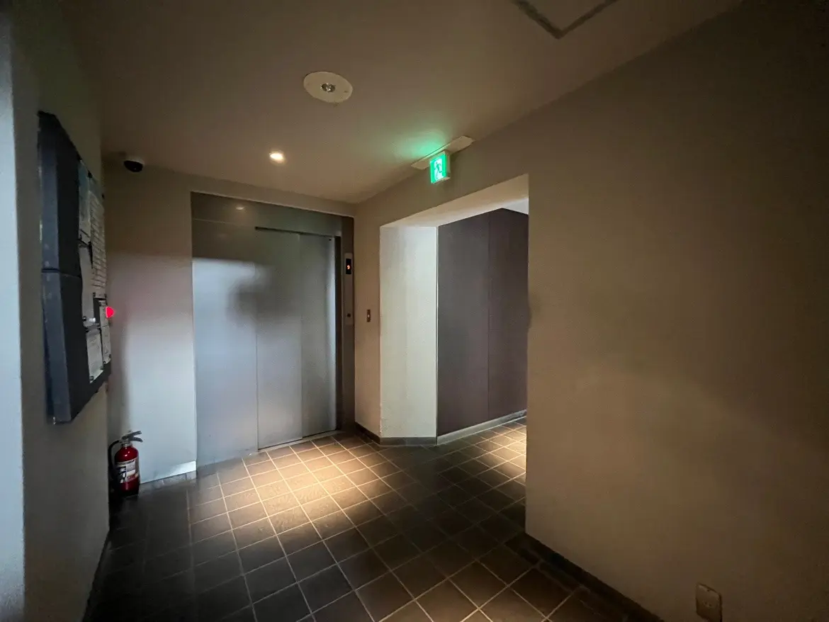 - シンプルでモダンなデザインのエレベーター前の廊下、床はタイル張りで火 extinguisherが置かれ、照明が控えめに配置されている様子