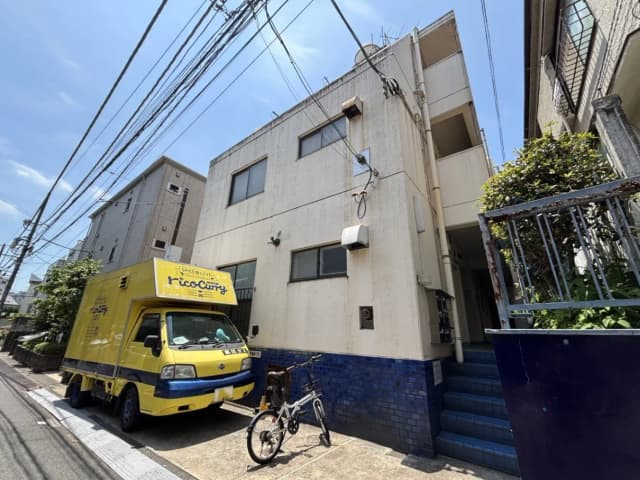 - 黄色い移動販売車が停まる、住宅街の閑静な通りと二階建ての建物、青いタイルの壁と急な階段がある景観